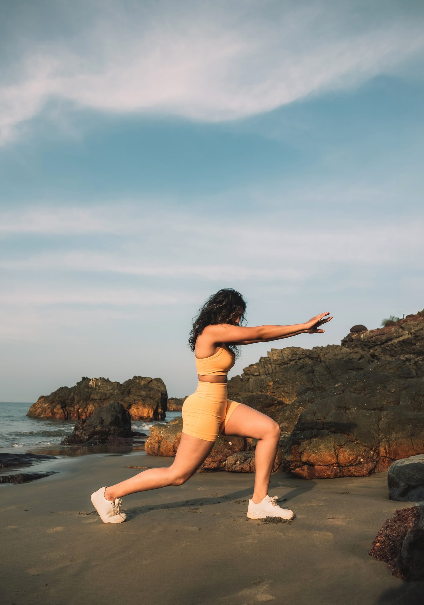 The model in yellow activewear holding a lunge on the beach, with her arms extended toward coastal rocks