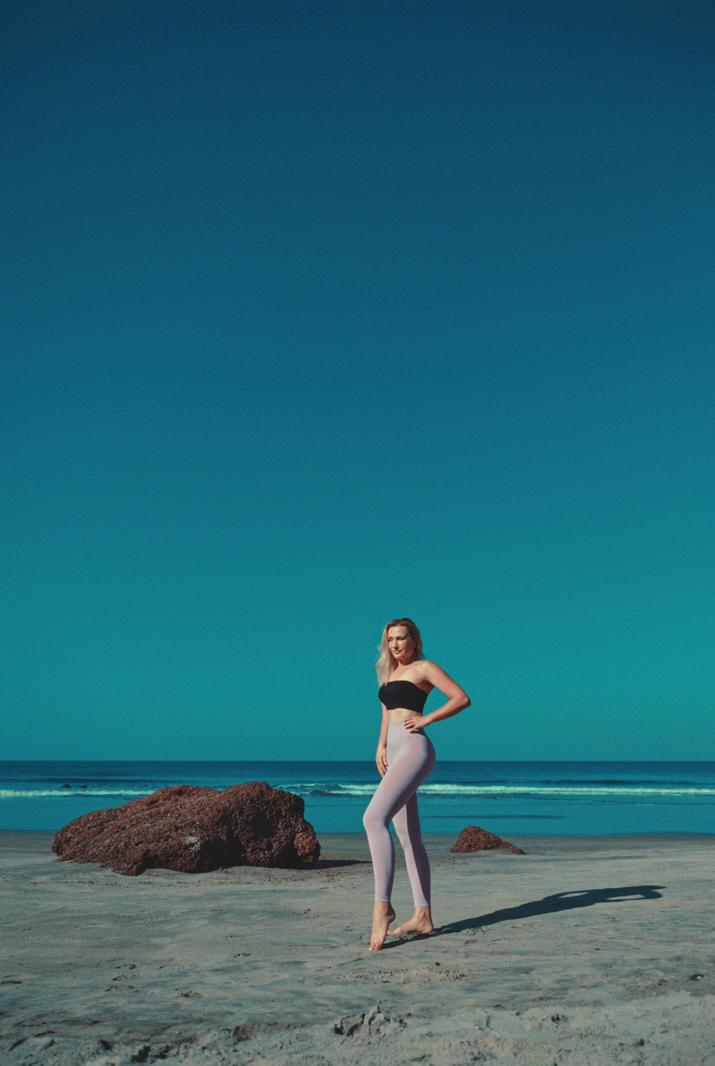 The model in a black bandeau top and lilac leggings standing on the beach beneath an expansive blue sky