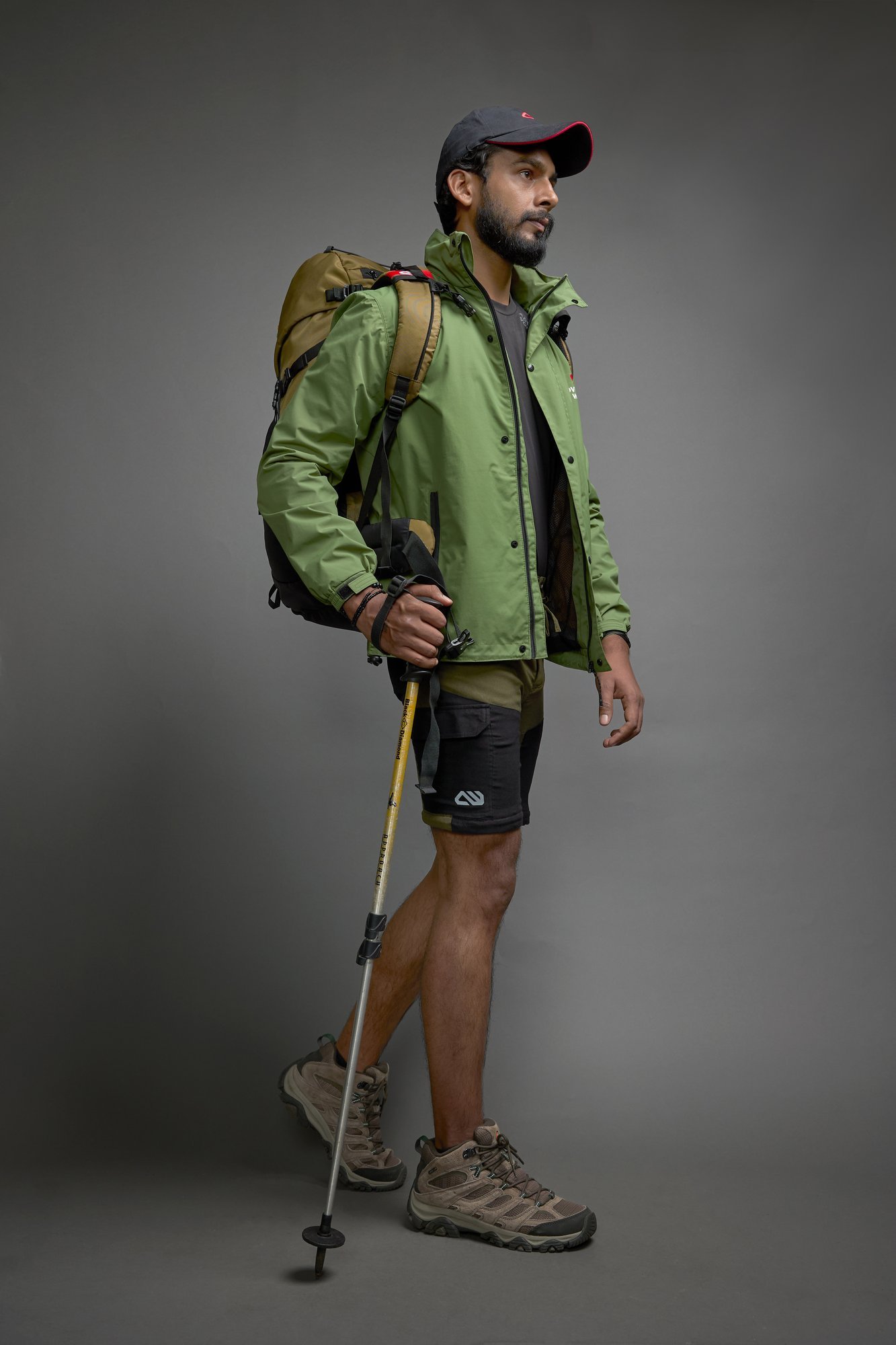 Hiker in a green jacket and cap walking forward with a trekking pole and backpack against a grey backdrop