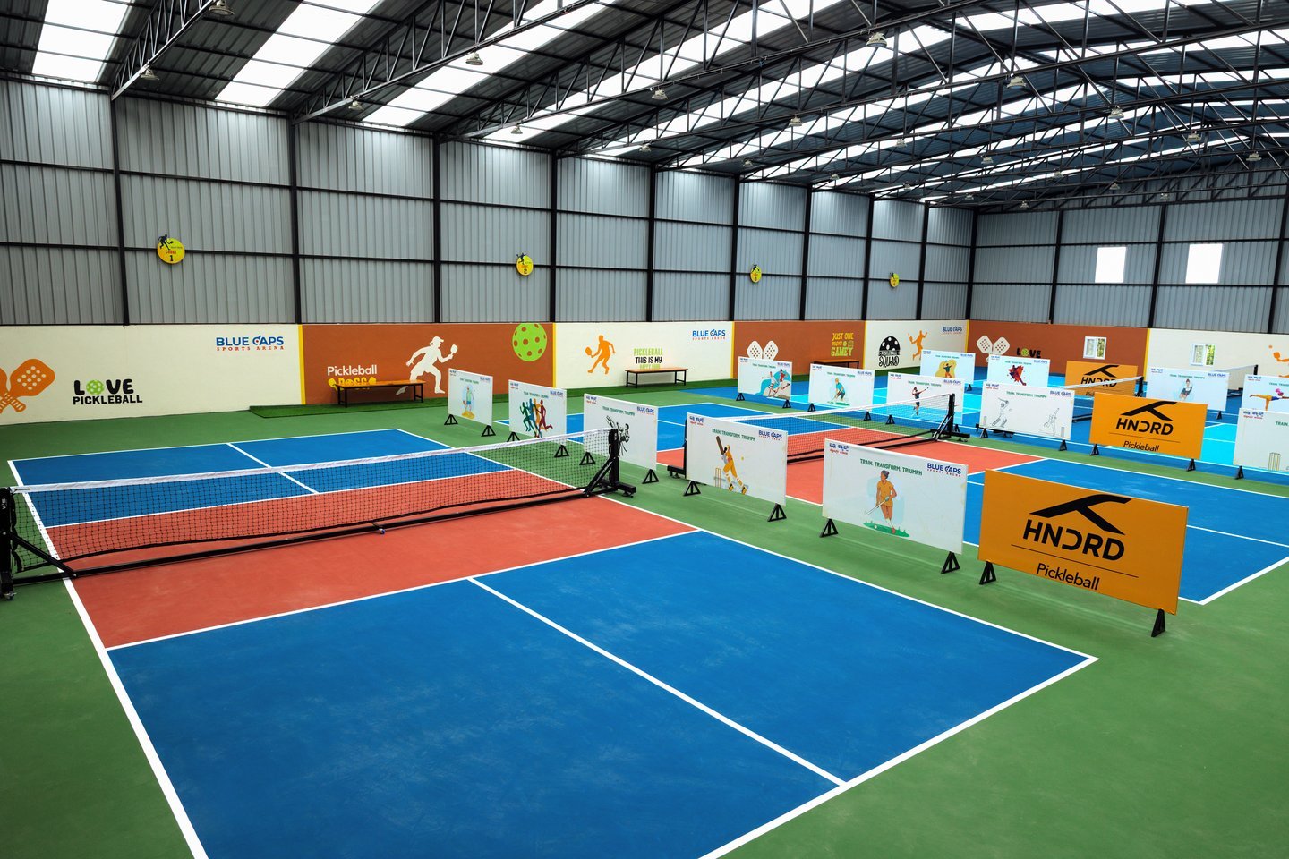 Indoor pickleball arena under a steel truss roof with skylights, blue and red courts in a row, player-graphic side panels and an orange HNDRD sign
