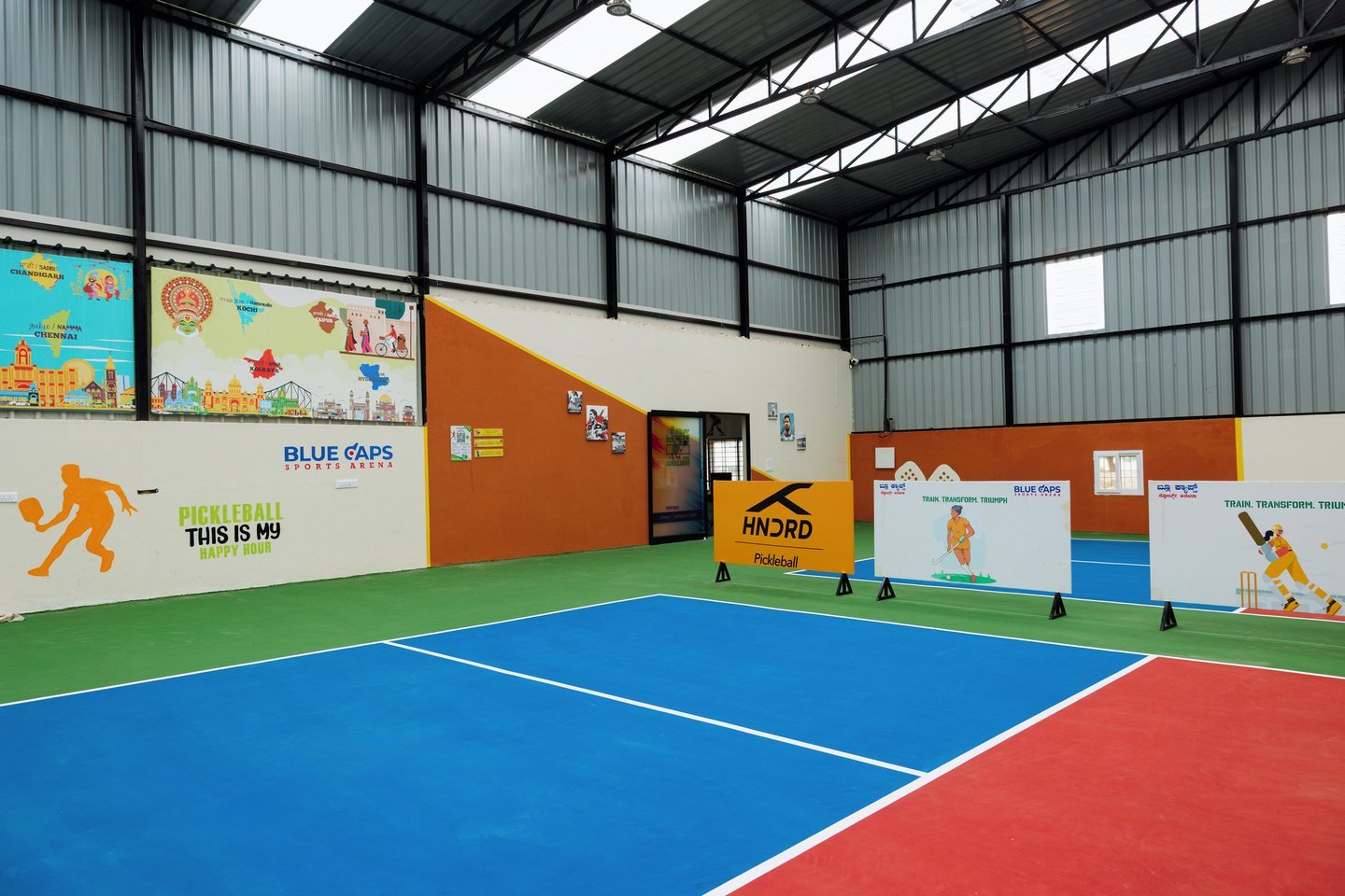 Corner of the arena with murals of pickleball players and a city skyline on orange and cream walls, a blue and red court in the foreground