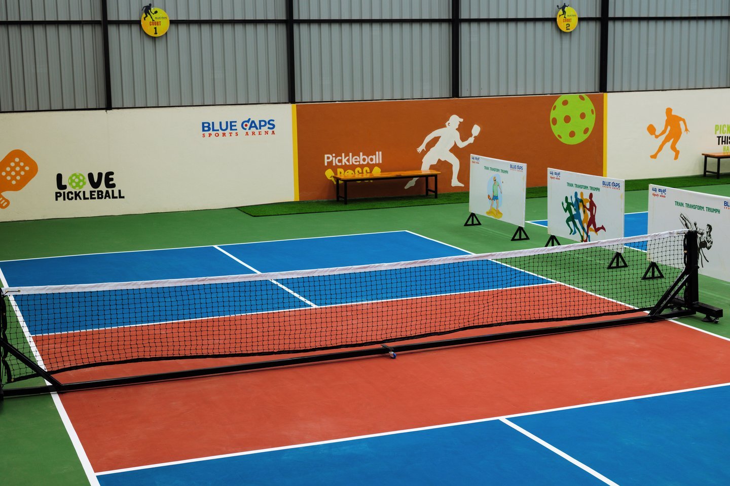 Pickleball net across a blue and red court, in front of a Love Pickleball wall, the Blue Caps logo and a player mural