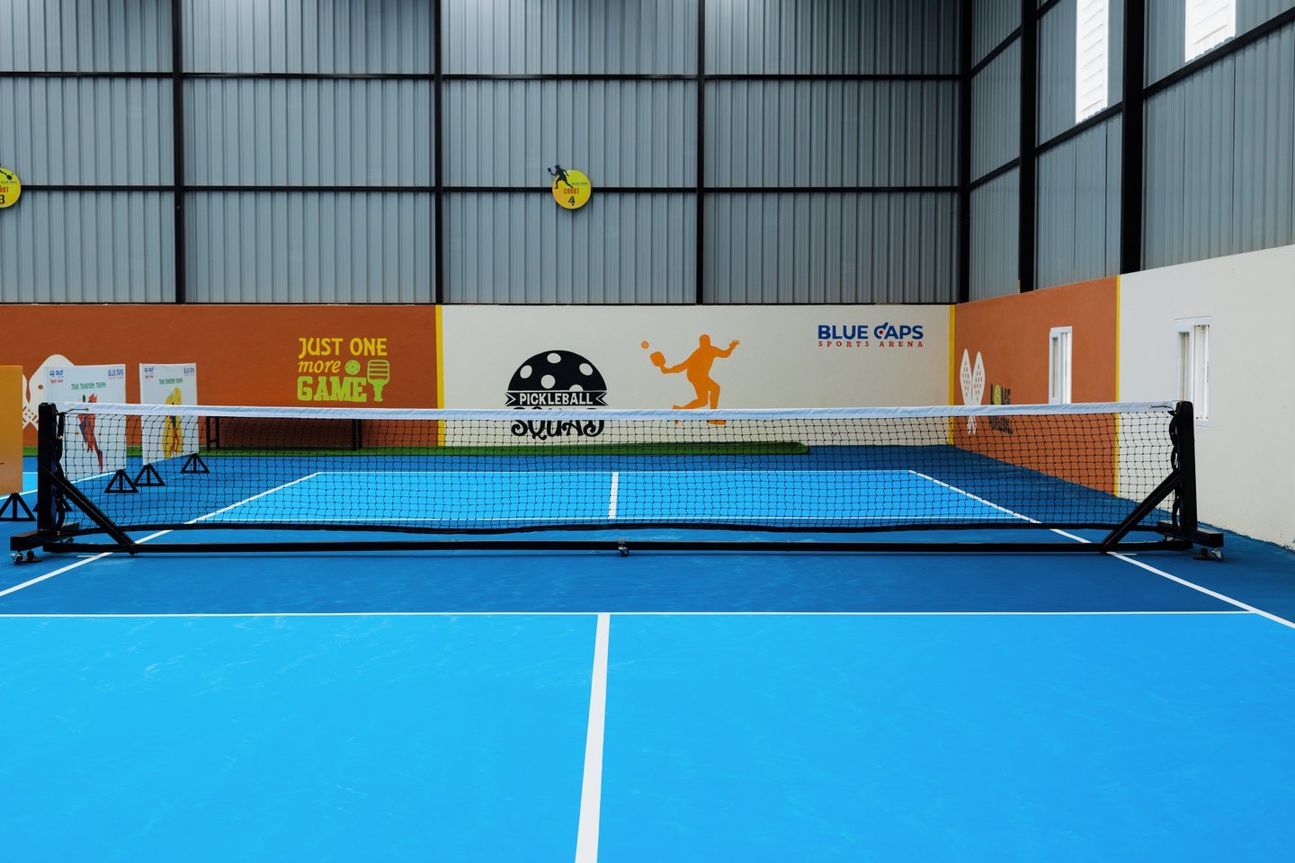 Straight-on view of a light blue court and net, facing a Just One More Game wall with the Blue Caps logo
