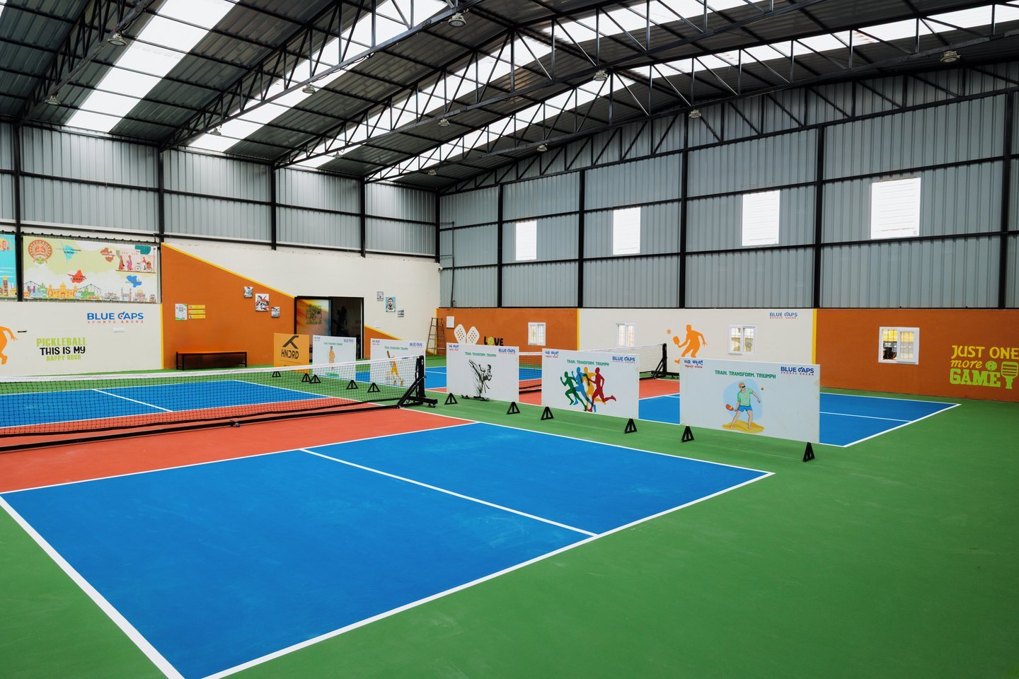 Wide view across the courts from a far corner, under the steel roof and high windows, with murals along the back wall