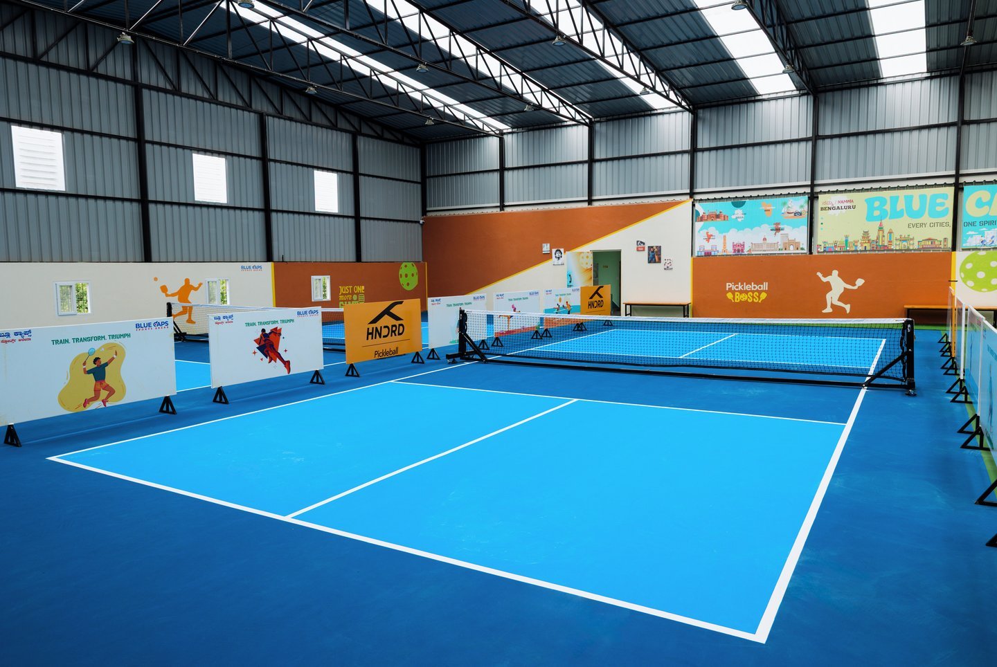 Light blue court and net in front of a colourful city mural, with player-graphic panels under the steel truss roof