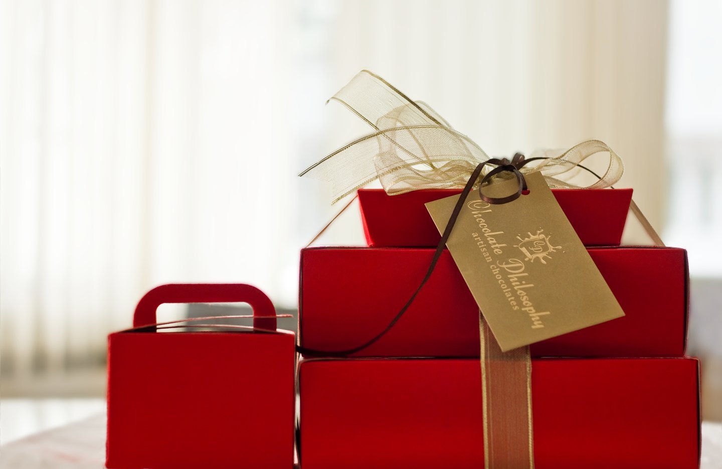 Stack of red Chocolate Philosophy gift boxes tied with a sheer ribbon and a gold gift tag, by a bright window