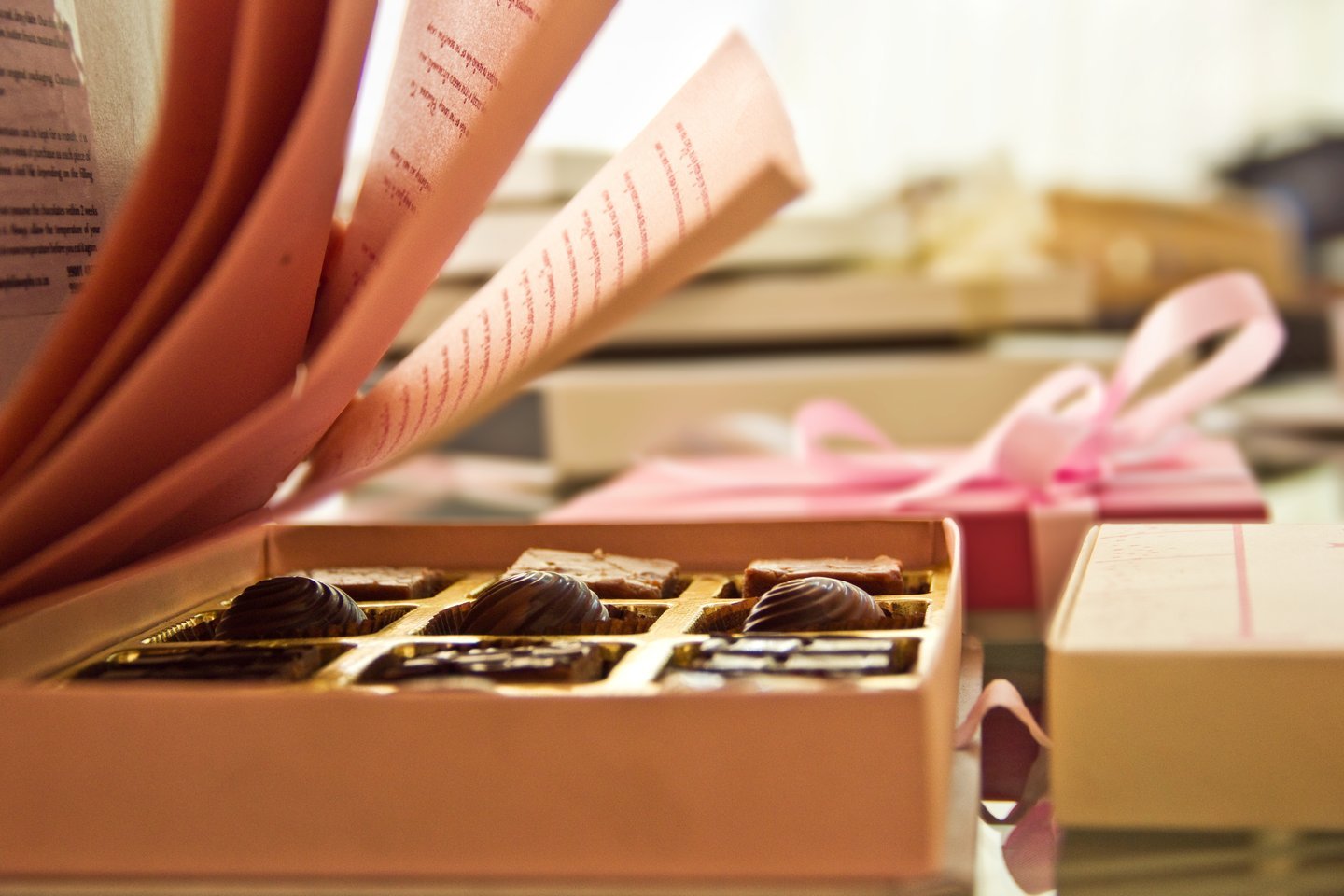 Open box of artisan chocolates in front of a book with fanned pink pages and a ribboned gift box
