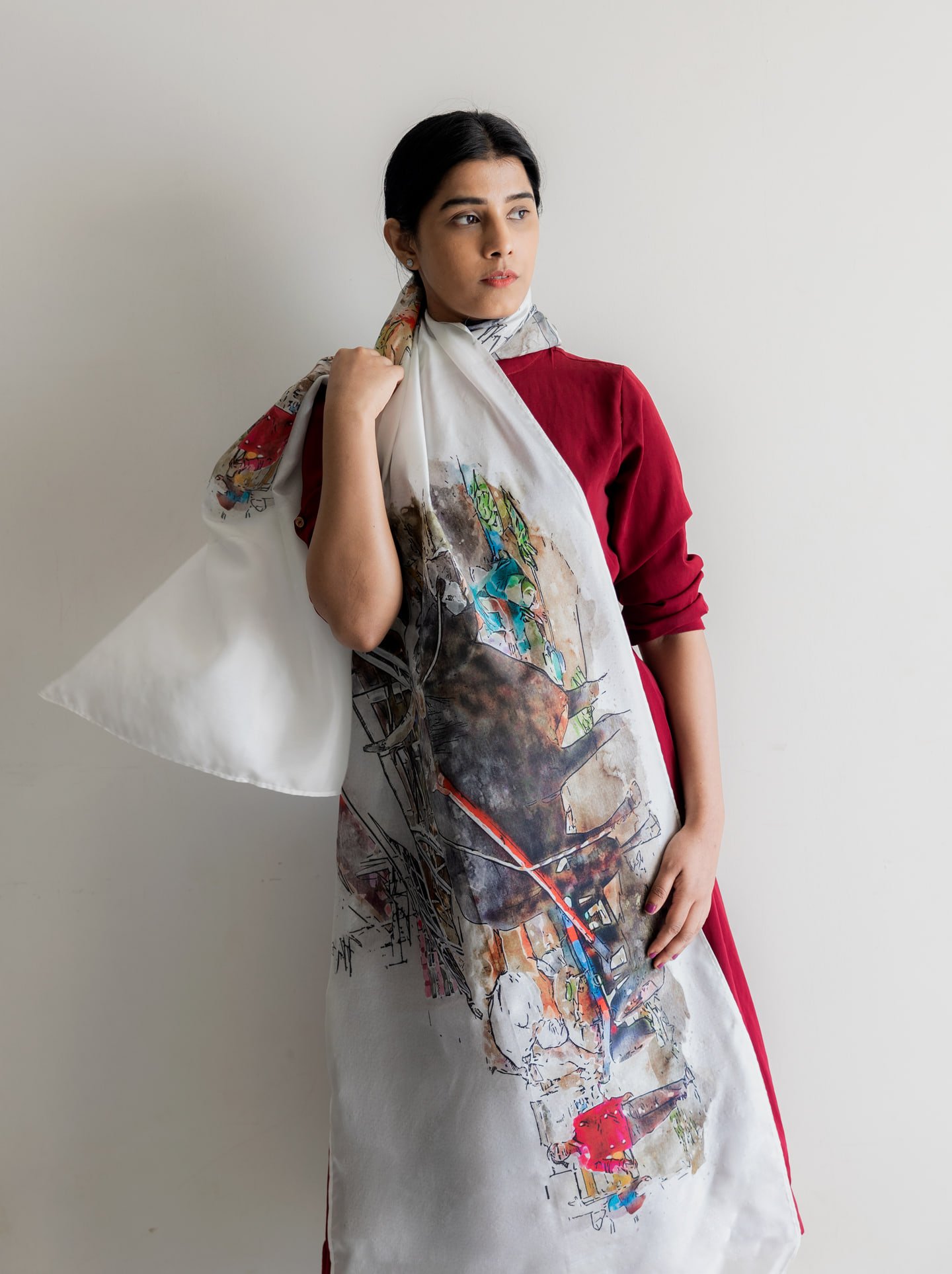 Model in a burgundy dress holding a flowing illustrated scarf, looking to the side against a pale wall