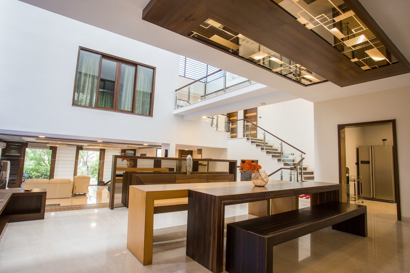 Double-height dining space with a long timber table and bench, orange flowers in a glass vase, a floating walnut ceiling with mirrored lighting and a glass-railed staircase