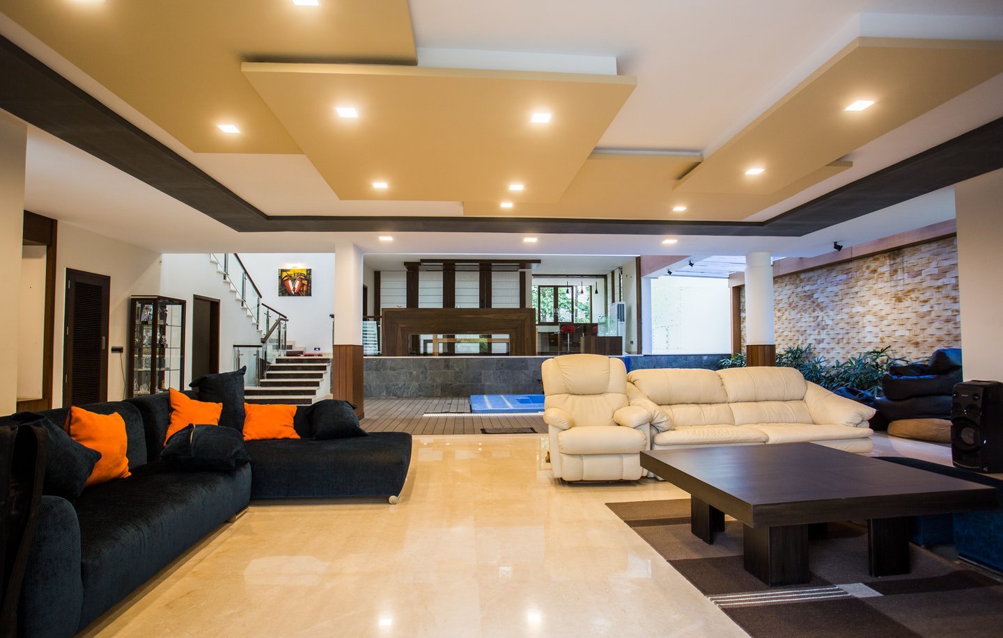 Living room with a black sectional sofa and orange cushions, cream recliners, a low timber table and a stepped ceiling with warm downlights