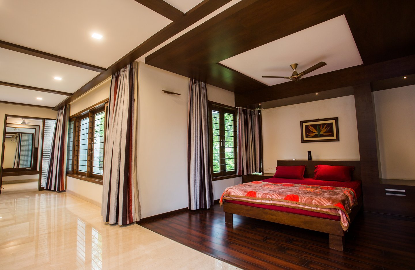 Bedroom with a red bedspread, a timber-framed ceiling with a fan, tall curtains and polished floors