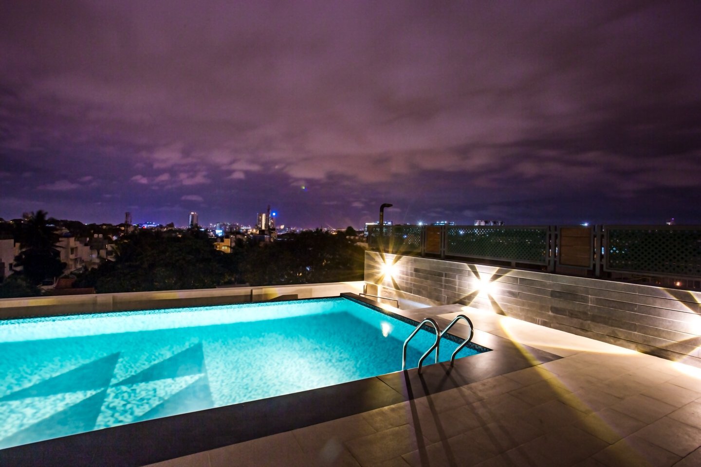 Rooftop pool lit turquoise at dusk under a purple sky, with the city lights on the horizon