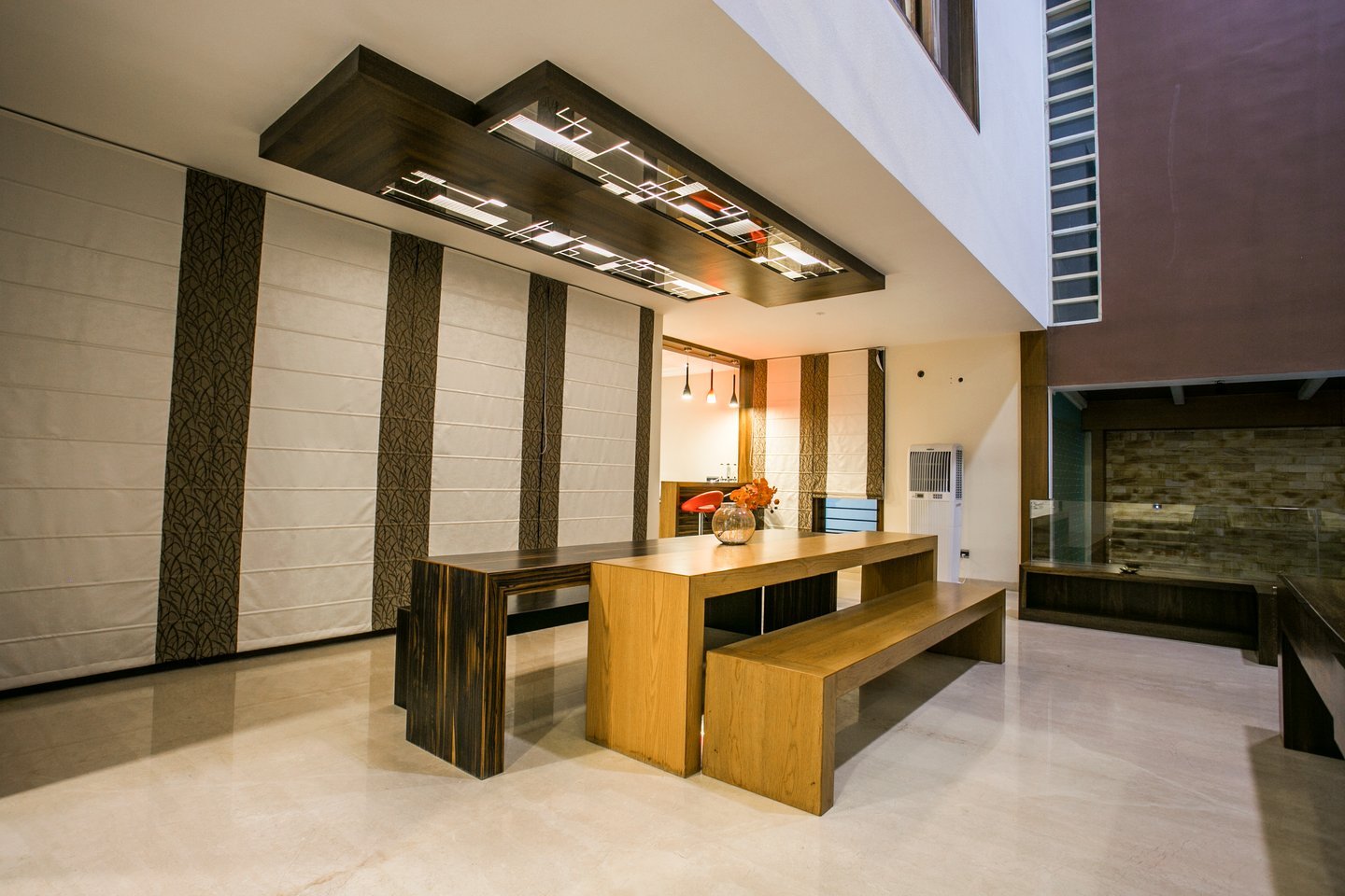 Dining area seen from the other side, with the long timber table and bench under the floating mirrored ceiling, roman blinds and a view through to the bar