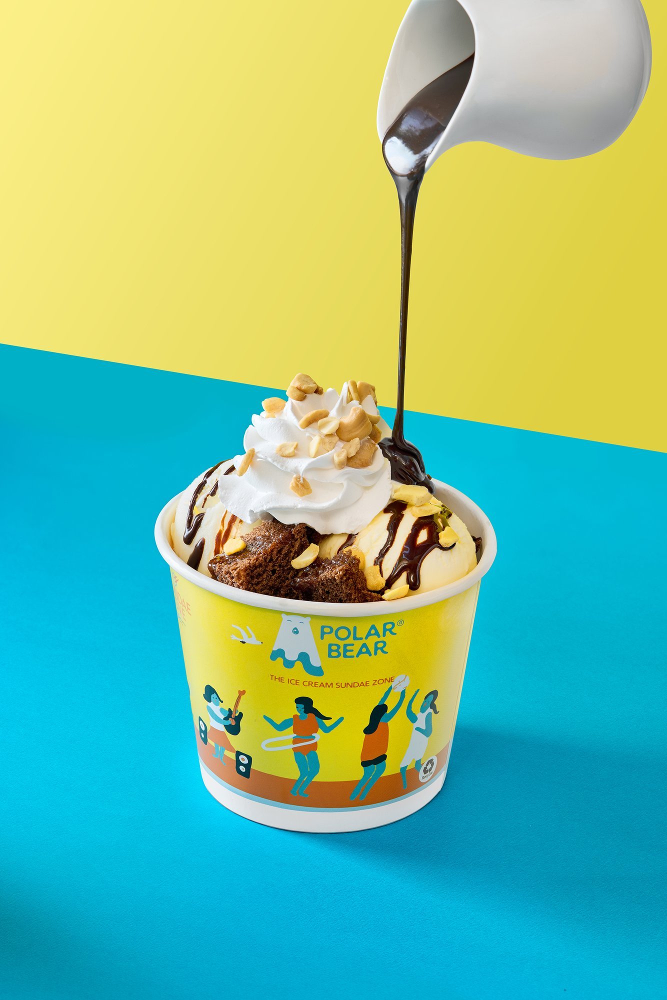 Chocolate sauce poured from a white jug onto a sundae in a Polar Bear cup, topped with cream and nuts, on a yellow and blue backdrop