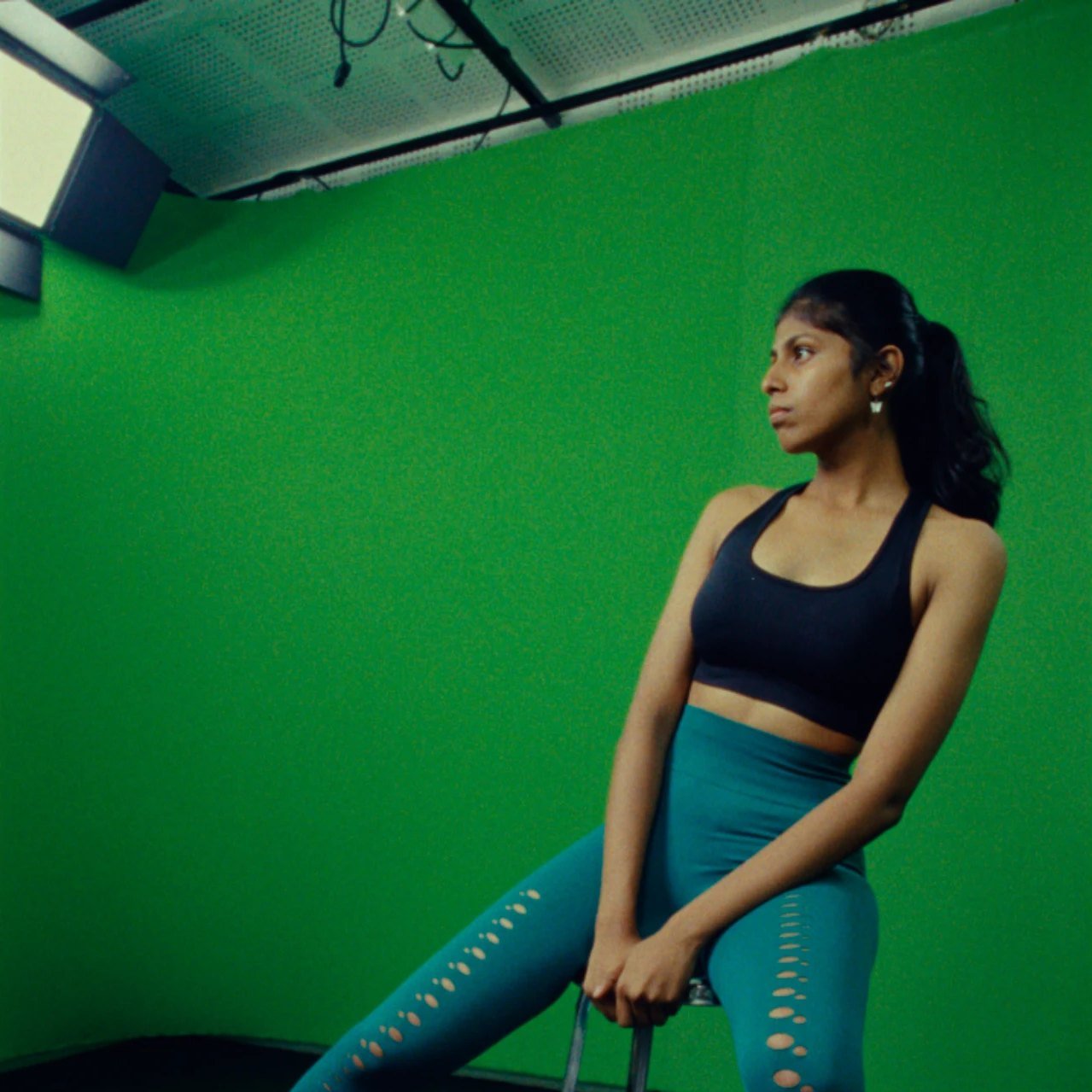 Model in a black sports top and teal leggings seated on a stool in a green studio, with a light panel and ceiling rig above her