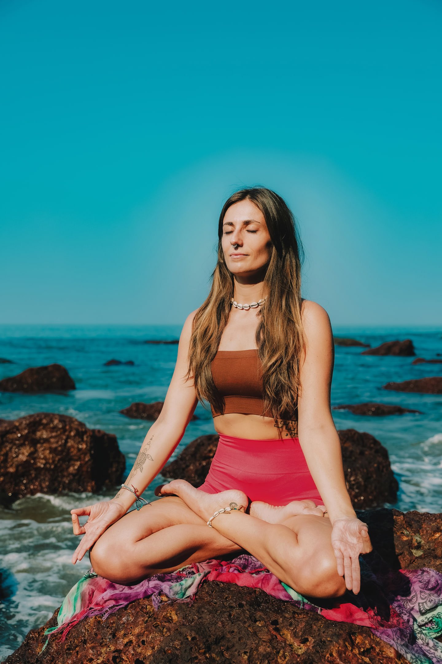 The model seated cross-legged on coastal rocks with eyes closed, wearing a brown top and pink shorts